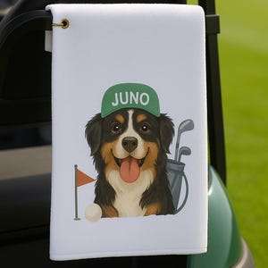 May include: A white golf towel with a cartoon Bernese Mountain Dog wearing a green baseball cap with the word 