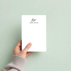 Può includere: Un blocco note rettangolare bianco con le iniziali js stampate in nero in corsivo, accentate da una piccola illustrazione di una foglia di eucalipto verde, con il nome JANE SMITH stampato in lettere maiuscole nere subito sotto.