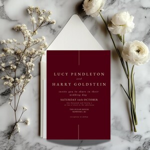 Puede incluir: Una invitación de boda burdeos con texto blanco, con los nombres "LUCY PENDLETON y HARRY GOLDSTEIN". La invitación incluye la fecha "SÁBADO 16 DE OCTUBRE" y detalles del evento. La invitación está sobre un fondo de mármol, con flores blancas y un sobre.