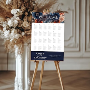 May include: A wedding seating chart on a wooden easel. The top of the chart has the word "WELCOME" above a floral design. The names "Emily and Zachary" are at the bottom, with the wedding date and location.
