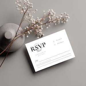May include: A white RSVP card with black text and a seashell graphic. The card includes lines for a response and dietary requirements. A gray vase with white flowers is in the background.