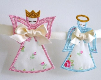 Puede incluir: Una guirnalda con dos ángeles bordados, uno en rosa y otro en azul, con una cinta blanca y tela floral.