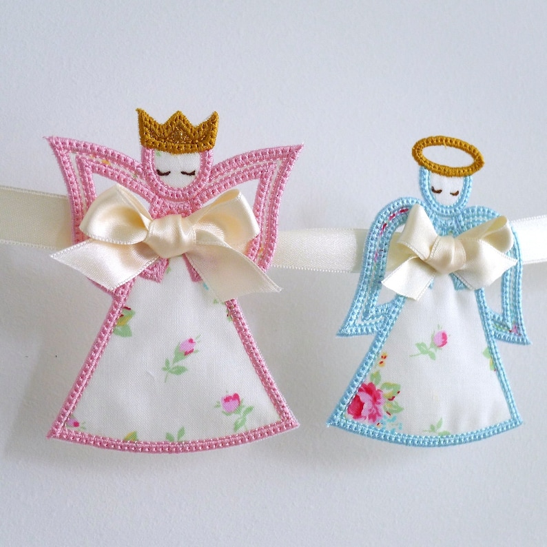 May include: A garland with two embroidered angels, one in pink and one in blue, with a white ribbon and floral fabric.