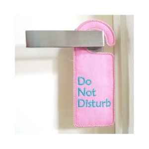 Könnte beinhalten: Ein rosa Türschild mit dem Text "Bitte nicht stören" in türkisblauer Stickerei.