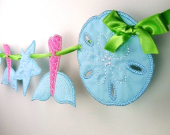 May include: A blue and pink fabric garland with a mermaid tail, starfish, and seashells. The garland is strung with a green ribbon and has a bow at the end.