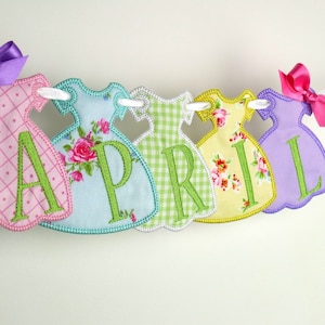 May include: A banner with the word "APRIL" spelled out in green letters on a string of six colourful fabric dresses. The dresses are pink, blue, green, yellow, and purple.