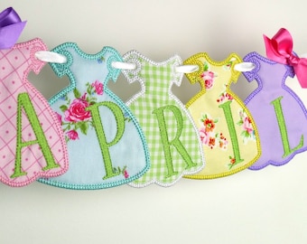 May include: A banner with the word "APRIL" spelled out in green letters on a string of six colorful fabric dresses. The dresses are pink, blue, green, yellow, and purple.