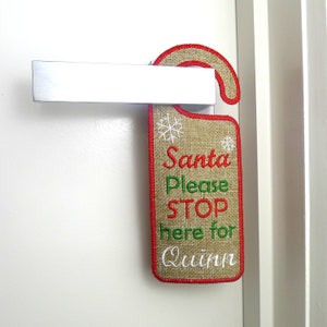 Könnte beinhalten: Ein Türhänger aus Jute mit roter und grüner Stickerei, auf dem "Santa Please Stop Here For Quinn" steht.