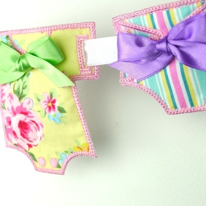 May include: A set of two fabric banners with pink trim. The banner on the left has a floral pattern and a green bow. The banner on the right has a striped pattern and a purple bow. Both banners are connected with a white ribbon.