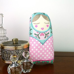 May include: A decorative, handmade Matryoshka doll with a pink polka-dot body and a teal and floral patterned head covering. The doll has closed eyes, pink cheeks, and a heart-shaped mouth. Other decorative items are in the background.