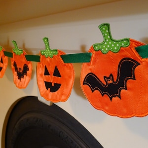 May include: A Halloween banner with orange felt pumpkins and a black felt bat. The pumpkins have green felt stems and stitched-on jack-o'-lantern faces. The banner is strung with green ribbon.