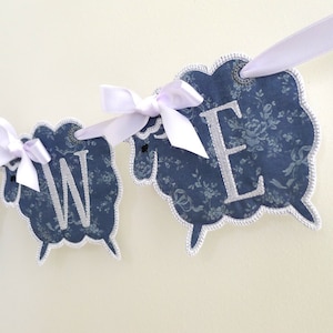 May include: A blue and white fabric banner with the letters "WE" in white embroidery. The banner is decorated with white bows and is strung together with white ribbon.