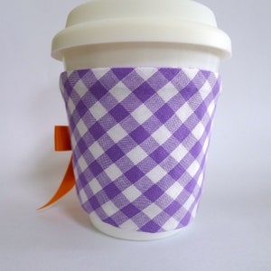 May include: A white to-go cup with a purple and white gingham fabric sleeve. An orange ribbon is tied around the cup.