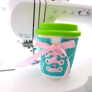 May include: A turquoise blue and white patterned coffee cup cozy with a pink ribbon tied in a bow. The cozy has white stitching that looks like shoelaces.