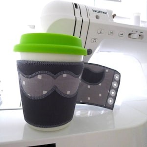 May include: A gray and white fabric coffee cup cozy with a stitched mustache design. The cozy is on a white surface next to a white sewing machine with the word "brother" on it.