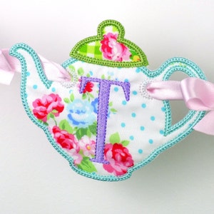 Op de afbeelding: Een turquoise theepot met een witte achtergrond en een bloemenprint. De theepot heeft een groen deksel met een bloemenpatroon en een paarse letter "T" geborduurd op de voorkant. De theepot heeft roze lintjes.