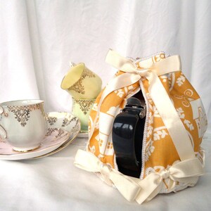 May include: A yellow and white teapot cozy with a white satin ribbon and bow. The cozy is decorated with a floral pattern and has a black handle.