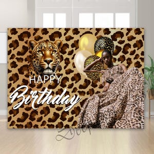 May include: A leopard print backdrop with a large leopard head and the words "Happy Birthday" in white script. A person wearing a leopard print dress is seated near gold and black balloons. The backdrop has a brown and tan spotted pattern.