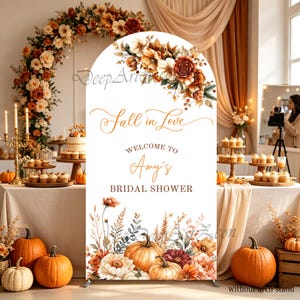 Könnte beinhalten: Ein weißes, bogenförmiges Hintergrundschild mit dem Text Fall in Love, Welcome to Amy's Bridal Shower. Das Design zeigt Aquarellillustrationen von orangefarbenen, braunen und cremefarbenen Blumen oben sowie Kürbisse mit Herbstlaub am unteren Rand.