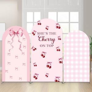May include: Three pink arched backdrop panels. One features a large bow and small bows, another has cherries and the text "She's the Cherry on Top", and the third has a pink gingham pattern. The panels are on silver stands.