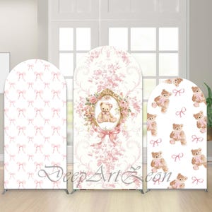 May include: Three arched backdrop stands in a soft pink theme. One has a repeating bow pattern, another a teddy bear in a floral frame, and the third has teddy bears and bows. The stands are on silver frames.