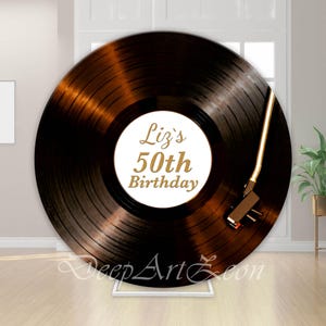 Pode incluir: Um grande disco de vinil castanho com um centro branco com o texto dourado "Liz's 50th Birthday". Um braço dourado estende-se lateralmente. O disco é exibido num suporte branco, evocando uma gira-discos vintage.
