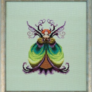 May include: Cross-stitch artwork of a fairy with a green and purple dress, red hair, and purple wings. The fairy is centered on a light green background, framed in a light wood-colored frame. The artwork is detailed and colorful.