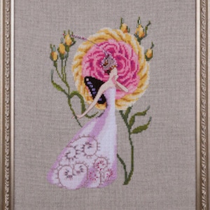 Puede incluir: Obra de arte de punto de cruz en un marco dorado. La imagen presenta a una mujer con un vestido rosa con una mariposa y una gran rosa amarilla y rosa. La obra de arte está detallada con hojas verdes y capullos amarillos. El fondo es beige neutro.