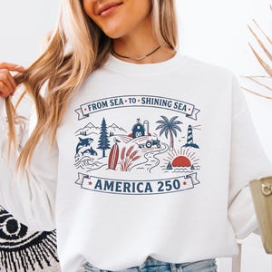 May include: White sweatshirt with a graphic design featuring a lighthouse, farm, and ocean scene with the text "FROM SEA TO SHINING SEA AMERICA 250". The sweatshirt is worn by a person.