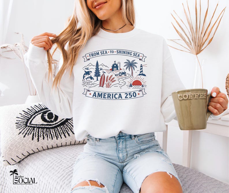 May include: White sweatshirt with a graphic design featuring a lighthouse, farm, and ocean scene with the text "FROM SEA TO SHINING SEA AMERICA 250". The sweatshirt is worn by a person.