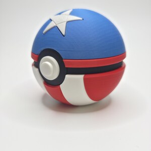 Peut inclure: Une Pok&eacute; Ball rouge, blanche et bleue avec une &eacute;toile blanche sur la moiti&eacute; sup&eacute;rieure bleue. La balle a une bande noire et un bouton blanc. Le design rappelle un th&egrave;me patriotique.