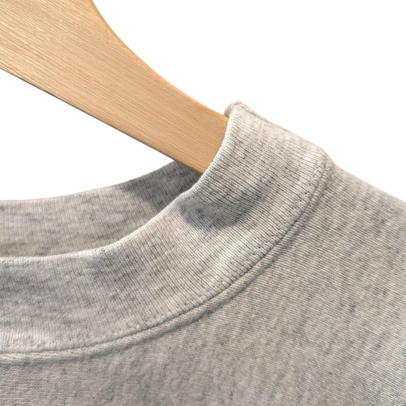 May include: Close-up of a heather gray crew neck sweatshirt hanging on a wooden hanger. The sweatshirt has a ribbed collar and a soft, textured appearance. The background is plain white.