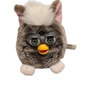 May include: A vintage Furby plush toy with a gray and brown speckled body, white fur on top, and pink-lined ears. It has large, expressive green eyes, a yellow beak, and white feet. The toy has a tag that reads "TIGER".