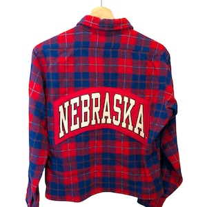 May include: A red, blue, and white plaid flannel shirt with the word "NEBRASKA" in white block letters across the back. The shirt is hanging on a wooden hanger.