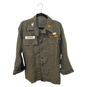 Op de afbeelding: Olijfgroene Amerikaanse legerjas met knoopsluiting, twee borstzakken en patches. De naam "MANTZ" staat op een naamplaatje. Patches zijn onder meer een gouden ster en een US Army patch.