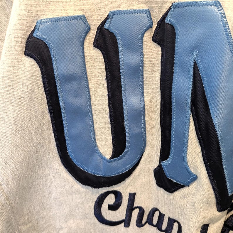 May include: Close-up of a light gray sweatshirt with the letters "UN" in light blue with a black outline. The word "Chan" is stitched in navy blue below the letters.