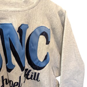 May include: A gray sweatshirt with the letters "NC" in light blue and navy blue, with the words "School Hill" below. The sweatshirt is made of a soft, textured material.