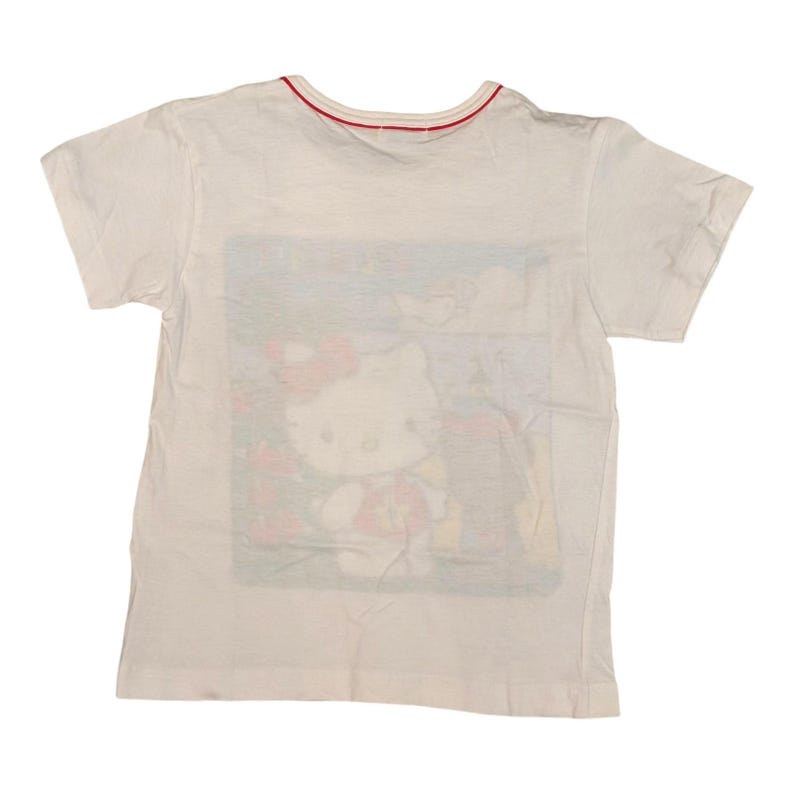 以下が含まれることがあります： ハローキティのグラフィックが特徴の白いTシャツ。Tシャツは半袖で、首周りには赤いトリムが付いています。グラフィックは赤いリボンと花柄の背景を持つハローキティを示しています。子供服。