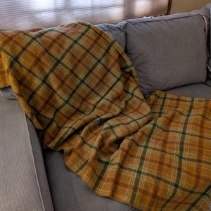May include: A rectangular throw blanket featuring a plaid pattern in shades of mustard yellow, forest green, and burnt orange. The fabric has a soft, textured appearance with a classic crisscross design that covers the entire surface of the textile.