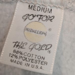 K&ouml;nnte beinhalten: Nahaufnahme eines wei&szlig;en Etiketts mit dem Wort "MEDIUM" oben. Das Etikett zeigt den Text "GO FOR" &uuml;ber einem kreisf&ouml;rmigen Logo und "THE GOLD" darunter. Es listet auch den Stoffgehalt als 88% Baumwolle und 12% Polyester auf und gibt "MADE IN U.S.A." an.
