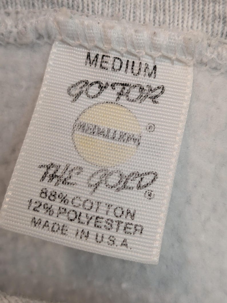 K&ouml;nnte beinhalten: Nahaufnahme eines wei&szlig;en Etiketts mit dem Wort "MEDIUM" oben. Das Etikett zeigt den Text "GO FOR" &uuml;ber einem kreisf&ouml;rmigen Logo und "THE GOLD" darunter. Es listet auch den Stoffgehalt als 88% Baumwolle und 12% Polyester auf und gibt "MADE IN U.S.A." an.