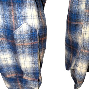 Pu&ograve; includere: Primo piano di una camicia di flanella a quadri blu, bianchi e marroni. La camicia presenta una tasca e maniche lunghe. Il tessuto ha un aspetto morbido e strutturato, con un classico motivo a quadri.