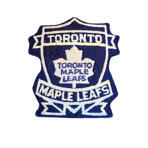 Puede incluir: Parche bordado azul marino y blanco con las palabras "TORONTO MAPLE LEAFS". El parche tiene forma de escudo con una hoja de arce en el centro. El texto "TORONTO" está en la parte superior y "MAPLE LEAFS" en la parte inferior.