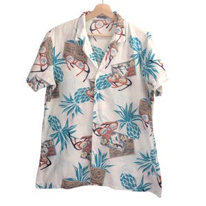 May include: White short-sleeve button-down shirt with a tropical print. The pattern includes teal pineapples, red-framed glasses, and vintage postcards. The shirt is hanging on a wooden hanger.