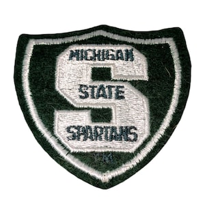 May include: A green and white embroidered patch in the shape of a shield. The patch features the words "MICHIGAN STATE SPARTANS" in white lettering, with a large white "S" in the center.