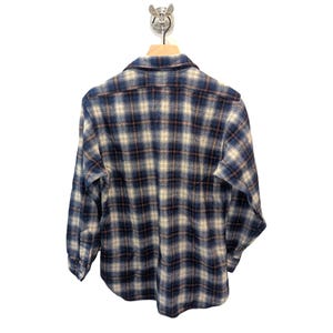 Pu&ograve; includere: Camicia di flanella a quadri blu, beige e marrone appesa a una gruccia di legno. La camicia a maniche lunghe &egrave; realizzata in un tessuto morbido e tessuto. Il motivo a quadri &egrave; un design classico.