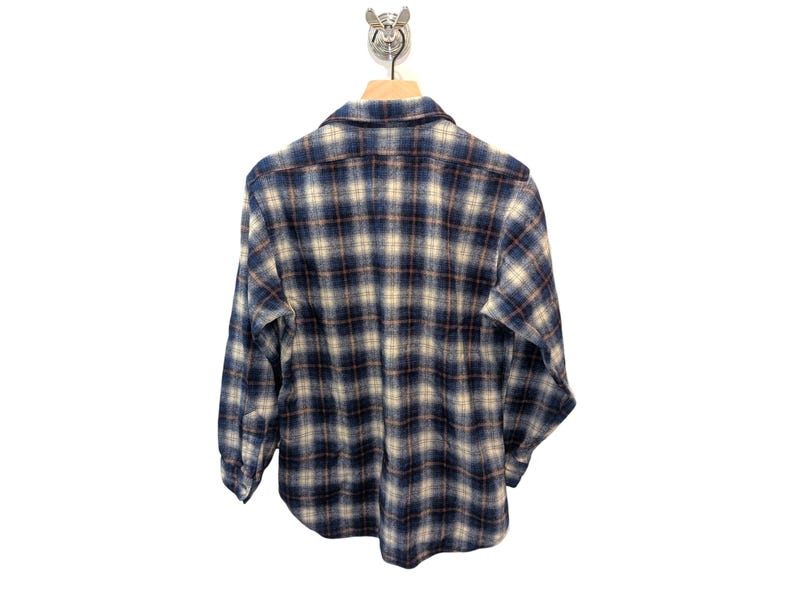 Pu&ograve; includere: Camicia di flanella a quadri blu, beige e marrone appesa a una gruccia di legno. La camicia a maniche lunghe &egrave; realizzata in un tessuto morbido e tessuto. Il motivo a quadri &egrave; un design classico.