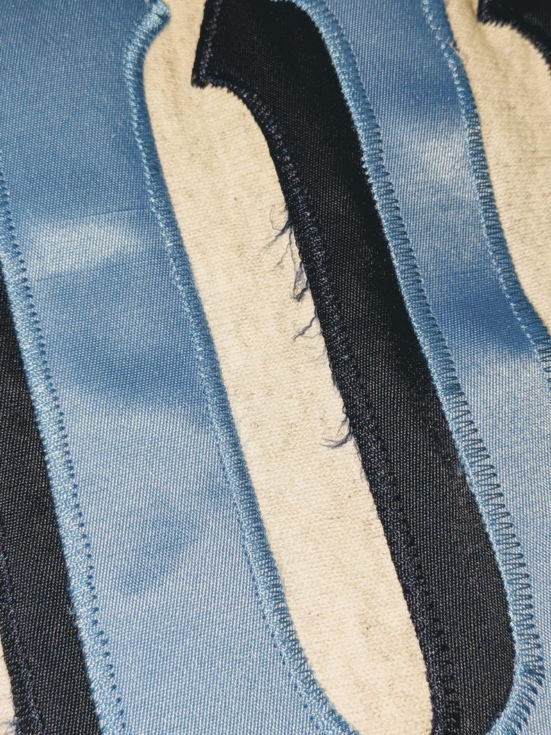 K&ouml;nnte beinhalten: Nahaufnahme eines Stoffes mit dem Buchstaben "N" im College-Stil. Der Buchstabe besteht aus hellblauem, schwarzem und cremefarbenem Stoff mit einer gen&auml;hten Bord&uuml;re. Der Stoff hat ein leicht abgenutztes Aussehen.