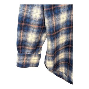 Pu&ograve; includere: Primo piano di una camicia di flanella a quadri blu, bianchi e pesca. Il tessuto &egrave; morbido e strutturato, con un motivo classico. La manica e il polsino sono visibili, suggerendo un design a maniche lunghe.