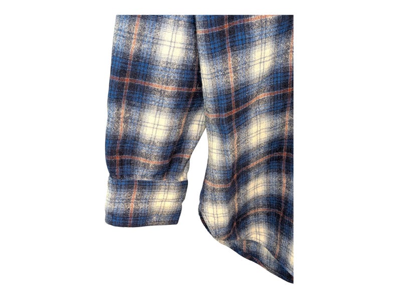 Pu&ograve; includere: Primo piano di una camicia di flanella a quadri blu, bianchi e pesca. Il tessuto &egrave; morbido e strutturato, con un motivo classico. La manica e il polsino sono visibili, suggerendo un design a maniche lunghe.